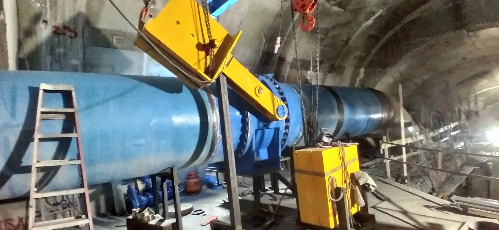Válvula montada en tubería azul dentro de túnel de hormigón, sostenida por viga de izaje amarilla y tecles