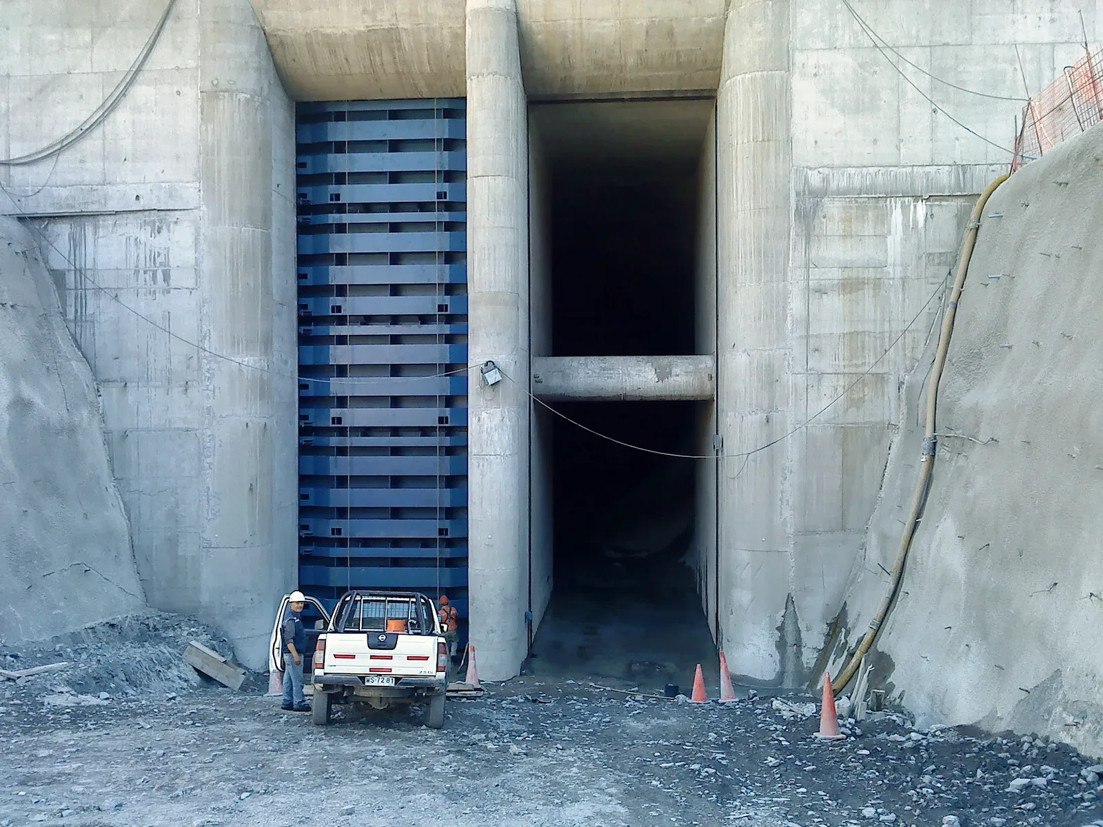 Compuerta plana azul de gran altura instalada en la boca de un túnel de hormigón, con camioneta y trabajador al pie